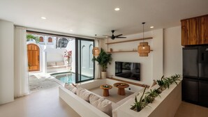 A modern living room with a sofa, two wooden side tables, a flat-screen TV, and a built-in kitchen area with a refrigerator. - Amal Villas by Bali Bliss Properties (Canggu)