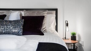 Ein ordentlich bezogenes Bett mit verschiedenen Kissen, ein Nachttisch mit Lampe und ein kleiner Topfpflanze.