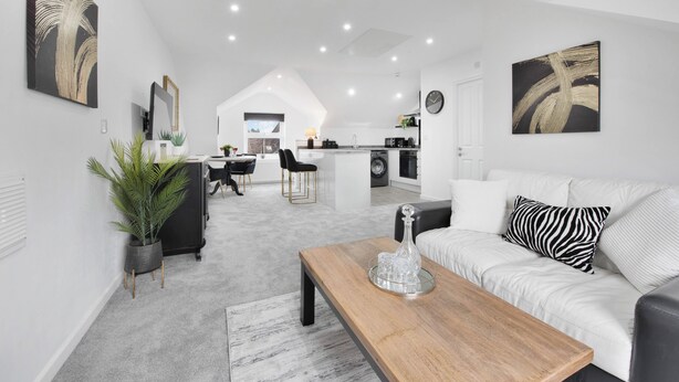 A modern living room with a wooden coffee table, a white sofa with a zebra-patterned pillow, and a dining area with a black chair and a plant.