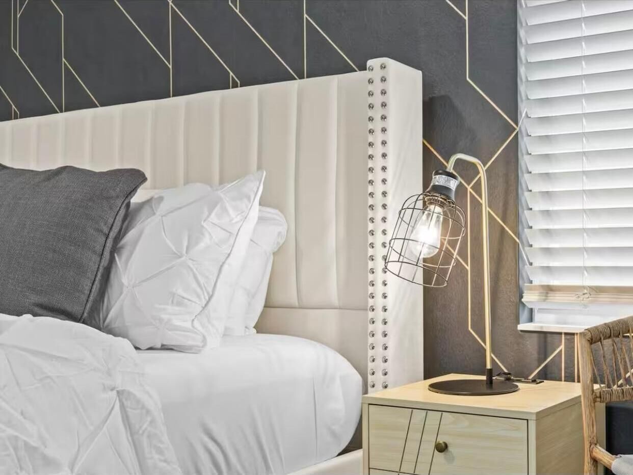 Un lit bien fait, avec une literie blanche, un oreiller gris et une table de chevet sur laquelle se trouve une lampe.
