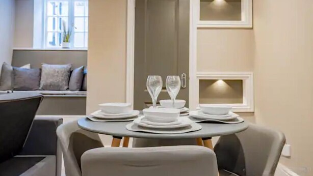 A dining area with a round table set for two, a sofa with cushions, and a window with a view. - WhiskAwayHomes Shaw Street Apartment 2 (Worcestershire)