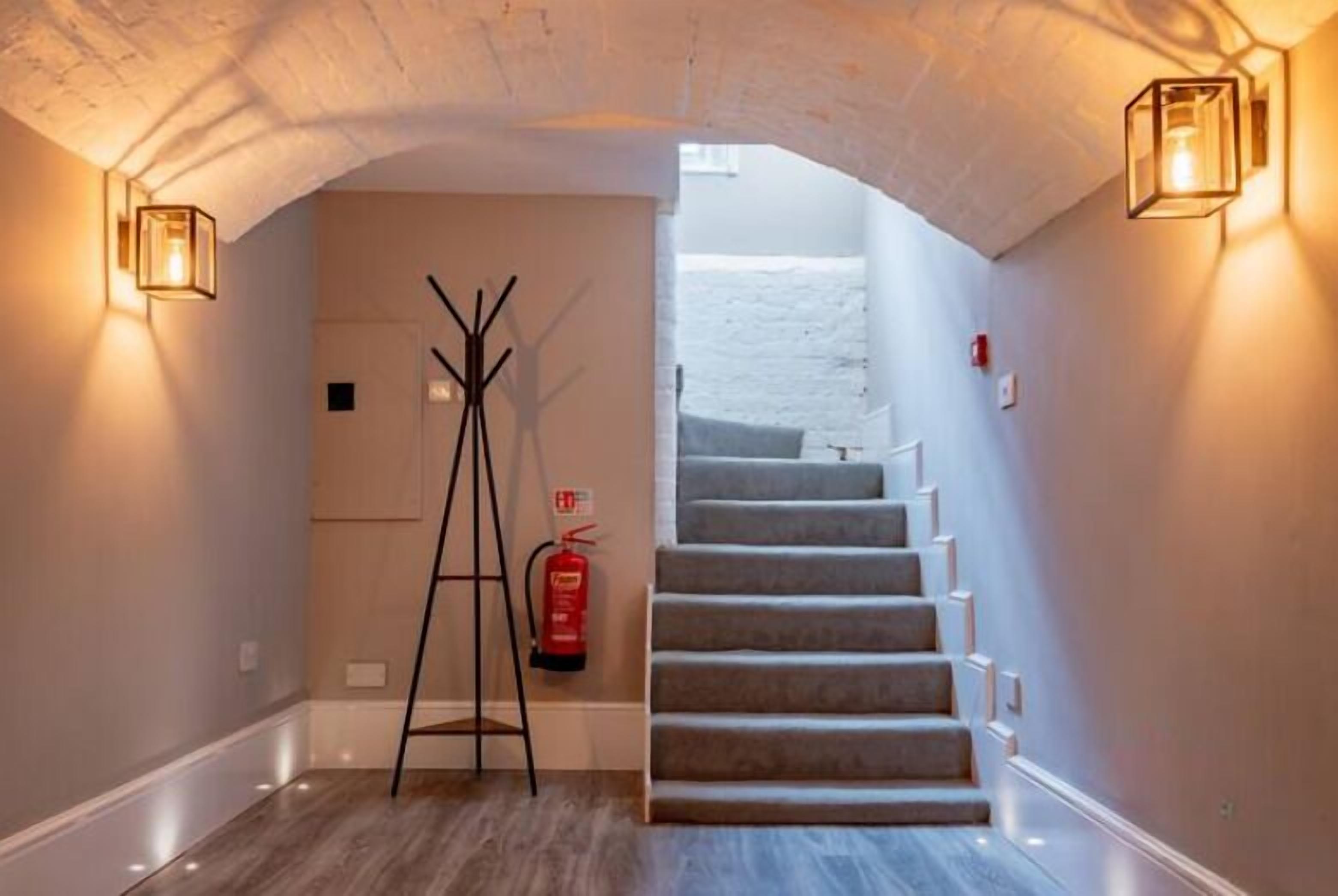 A staircase with a fire extinguisher and a ladder in a hallway with a white ceiling and wall sconces.