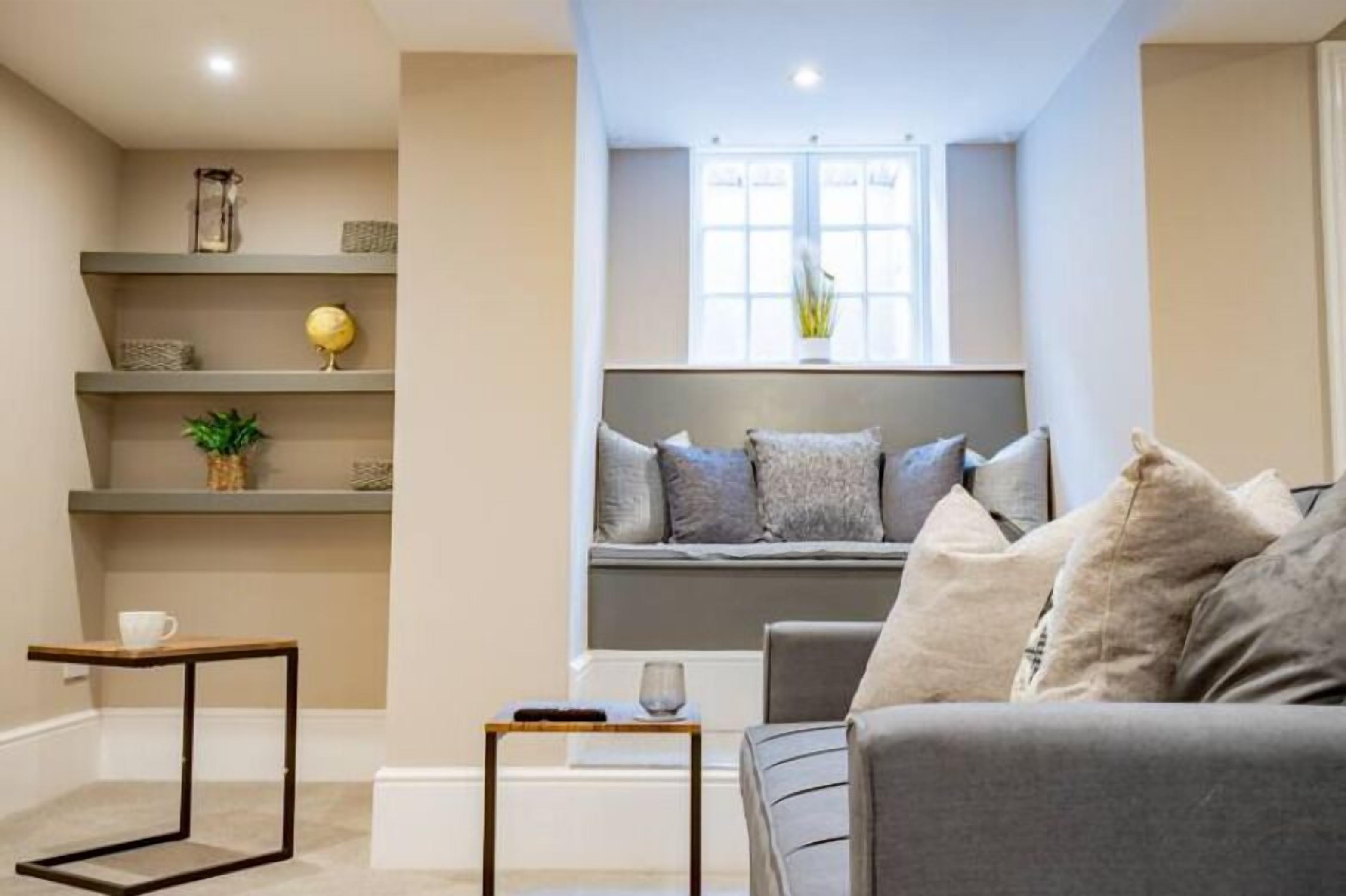 A modern living room with a grey sofa, a small table, and a window seat with cushions. Shelves with decorative items are visible.