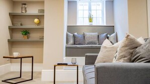 A modern living room with a grey sofa, a small table, and a window seat with cushions. Shelves with decorative items are visible. - WhiskAwayHomes Shaw Street Apartment 2 (Worcestershire)