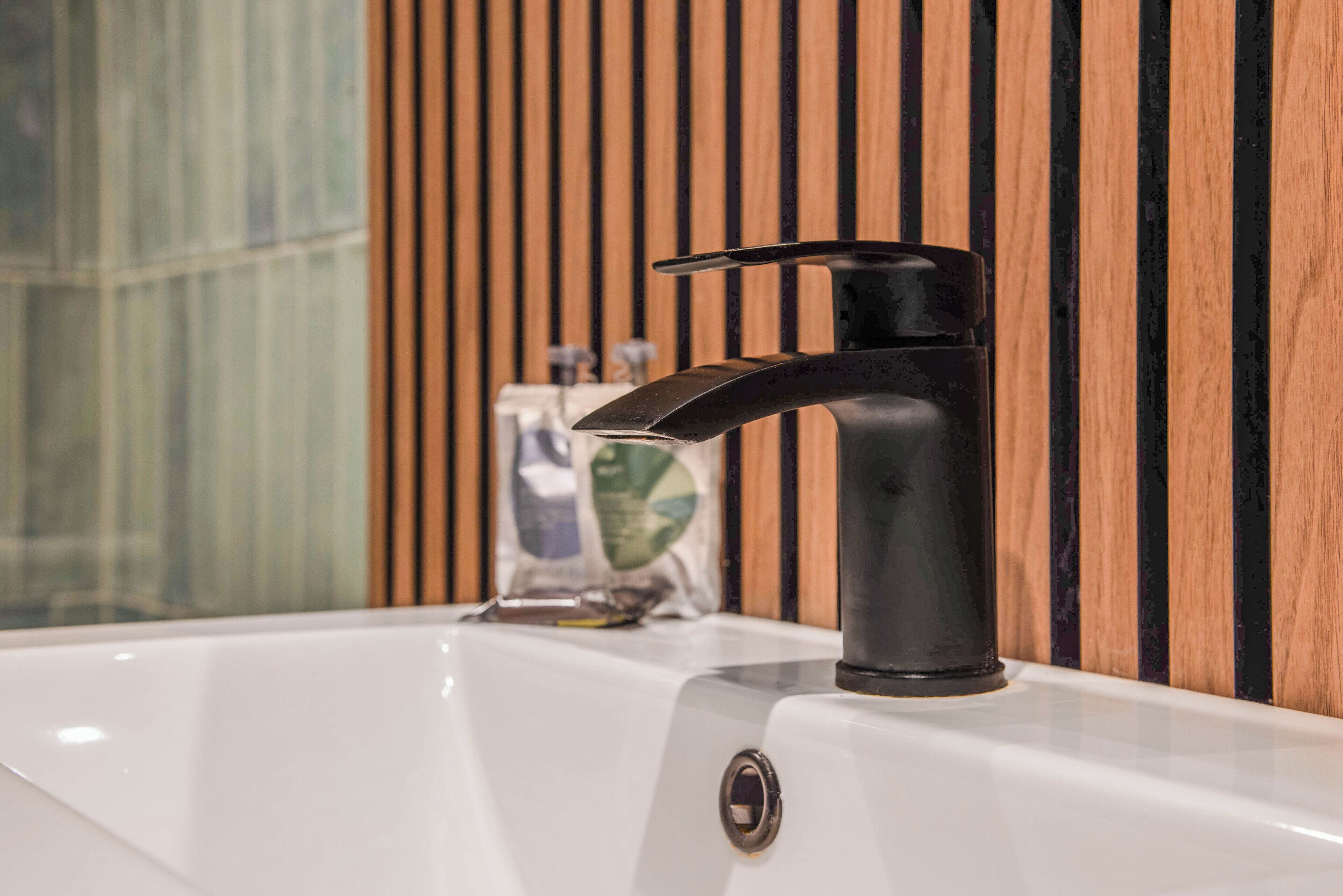 Een moderne badkamer met een strakke zwarte kraan, een witte wasbak en een wandpaneel van hout.