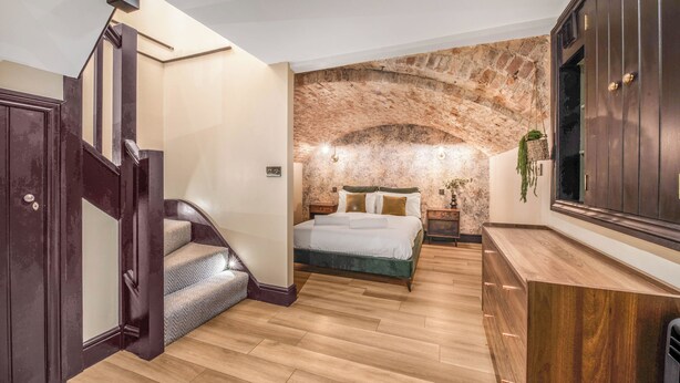 Un dormitorio con suelo de madera, una cama con cabecera verde, una escalera de madera oscura y un arco de pared de piedra.