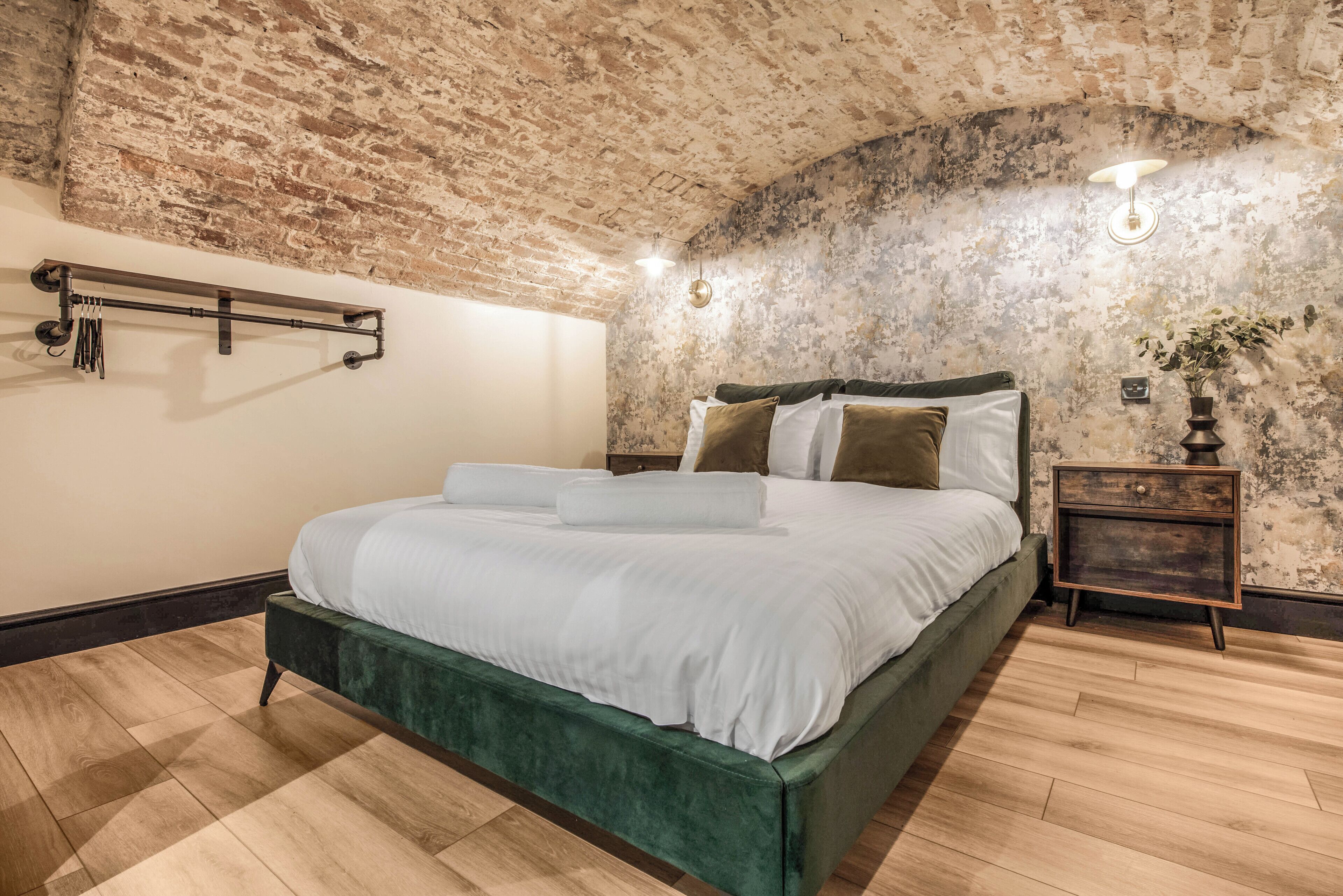 Un dormitorio con techo de ladrillo, una cama con ropa blanca, una mesita de noche de madera y una lámpara fijada a la pared.