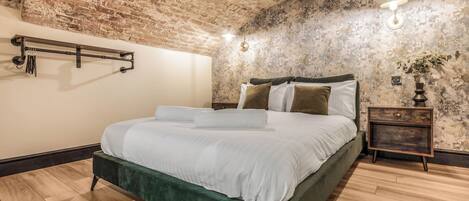Un dormitorio con techo de ladrillo, una cama con ropa blanca, una mesita de noche de madera y una lámpara fijada a la pared.