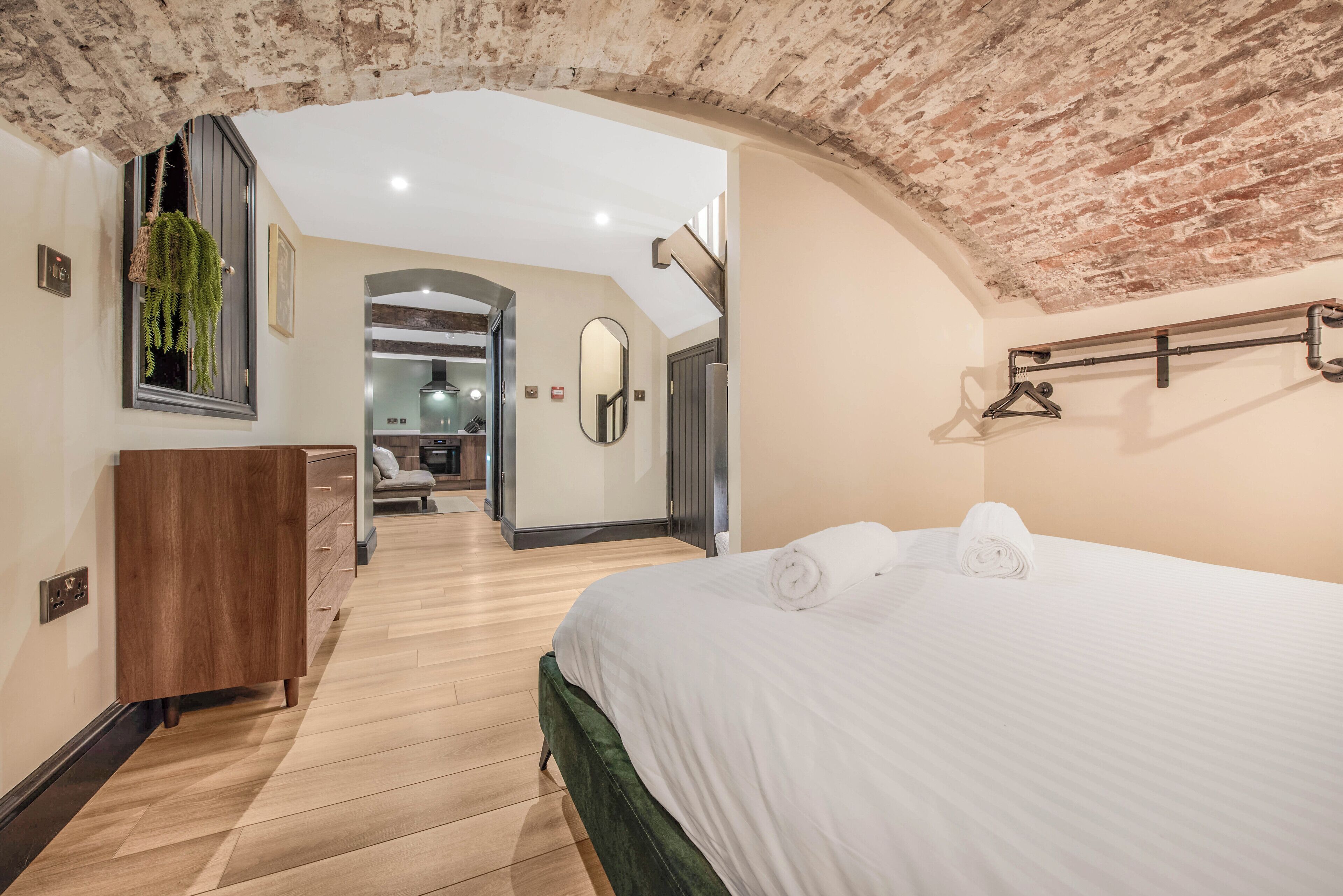 A hotel room with a wooden floor, a bed with white linens, a wooden dresser, a hanging plant, and a brick ceiling.
