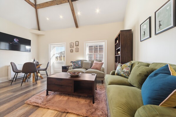 A living room with a wooden coffee table, a green sofa, a dining area with chairs, and a TV mounted on the wall. - WhiskAwayHomes Vinegar House No. 7 (Worcestershire)