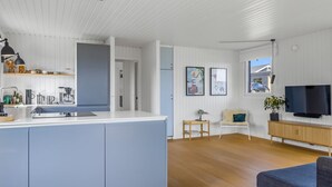 Una cocina moderna con una isla central, un área de comedor con un sofá y un televisor, y vistas al exterior a través de una ventana.