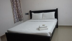 A neatly made bed with white linens and a folded towel, a dark wooden headboard, and a plain wall with a curtain. - Vasantharaja Villa Ariyankuppam (Puducherry)