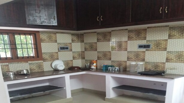 A kitchen with dark wooden cabinets, a tiled backsplash, and white countertops. There are two sinks, a stove, and various kitchen utensils. - Vasantharaja Villa Ariyankuppam (Puducherry)