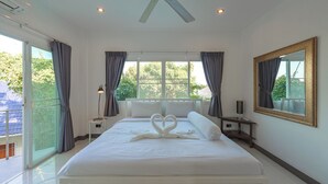 Ein geräumiges Schlafzimmer mit einem großen Bett, einem Nachttisch, einem Deckenventilator und Blick auf Grün hinter den Fenstern.