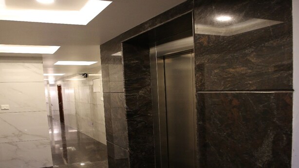 A modern hallway with a polished marble floor, a wall-mounted elevator, and recessed ceiling lighting. - Vasantharaja Inn (Puducherry)