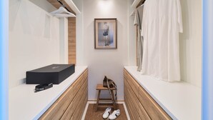 Ein schmaler begehbarer Kleiderschrank mit Holzboden, einem gerahmten Kunstwerk an der Wand, einer schwarzen Kiste, einer braunen Tasche und einem Paar Schuhe.