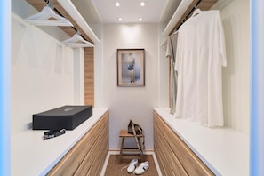 A narrow walk-in closet with wooden flooring, a framed artwork on the wall, a black box, a brown bag, and a pair of shoes.