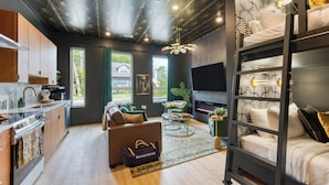 A modern living room with a sofa, coffee table, and a TV. There is a kitchen area with cabinets and a stove. The room has a unique ceiling design and large windows.