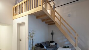 Et soveværelse med skråvægge, en trætrappe, en grå sofa, et lille hvidt bord og en potteplante.