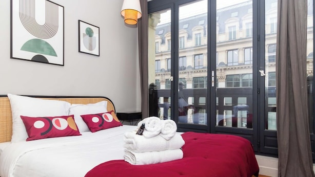 A hotel room with a large bed, a red bedspread, white bedding, and a view of buildings through a large window. - Superb Apartement 2 Bedrooms Center of Paris (Paris)