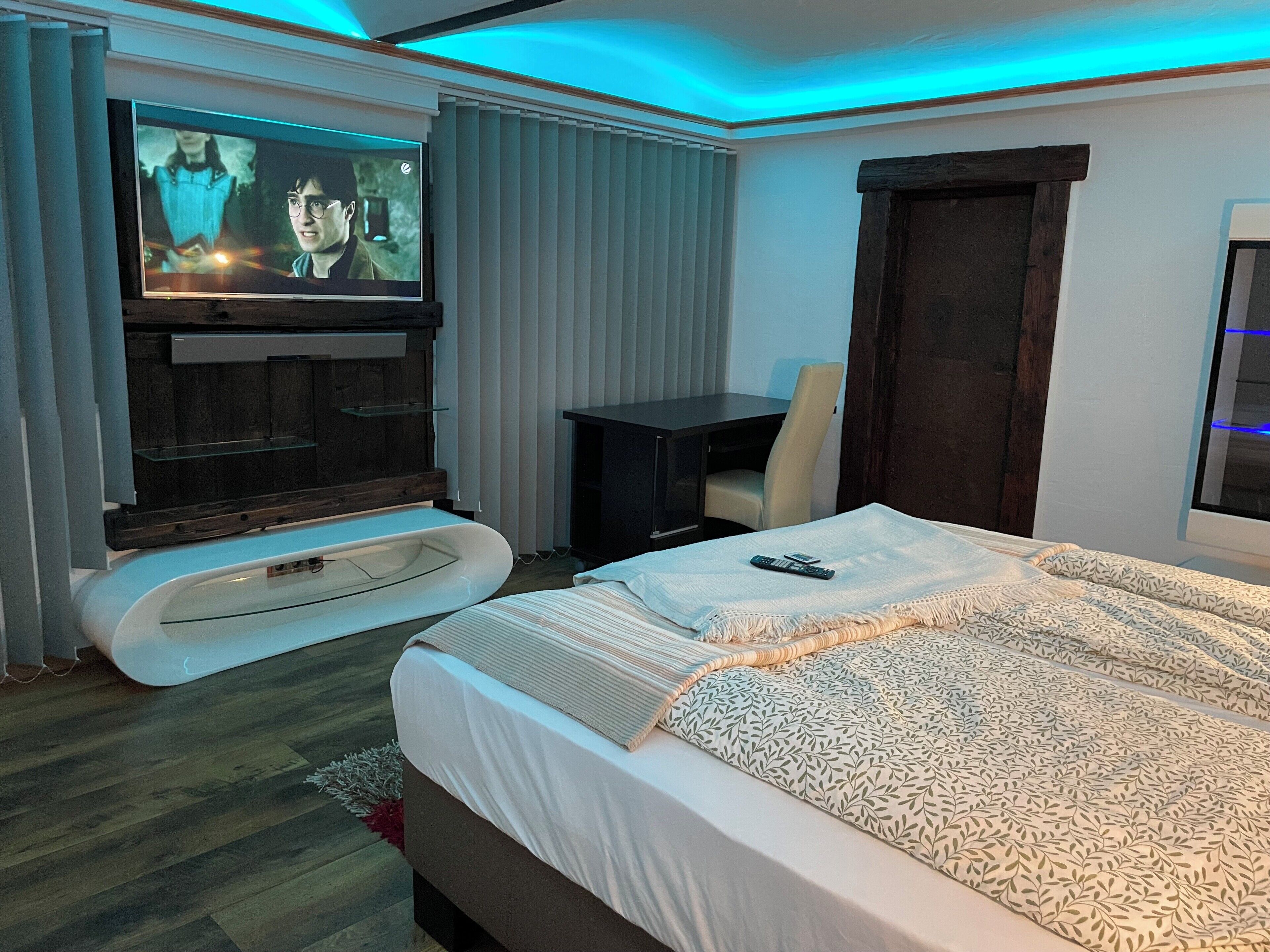 Une chambre d’hôtel moderne avec un grand lit, un bureau, un téléviseur fixé sur un panneau en bois et une porte en bois foncé.