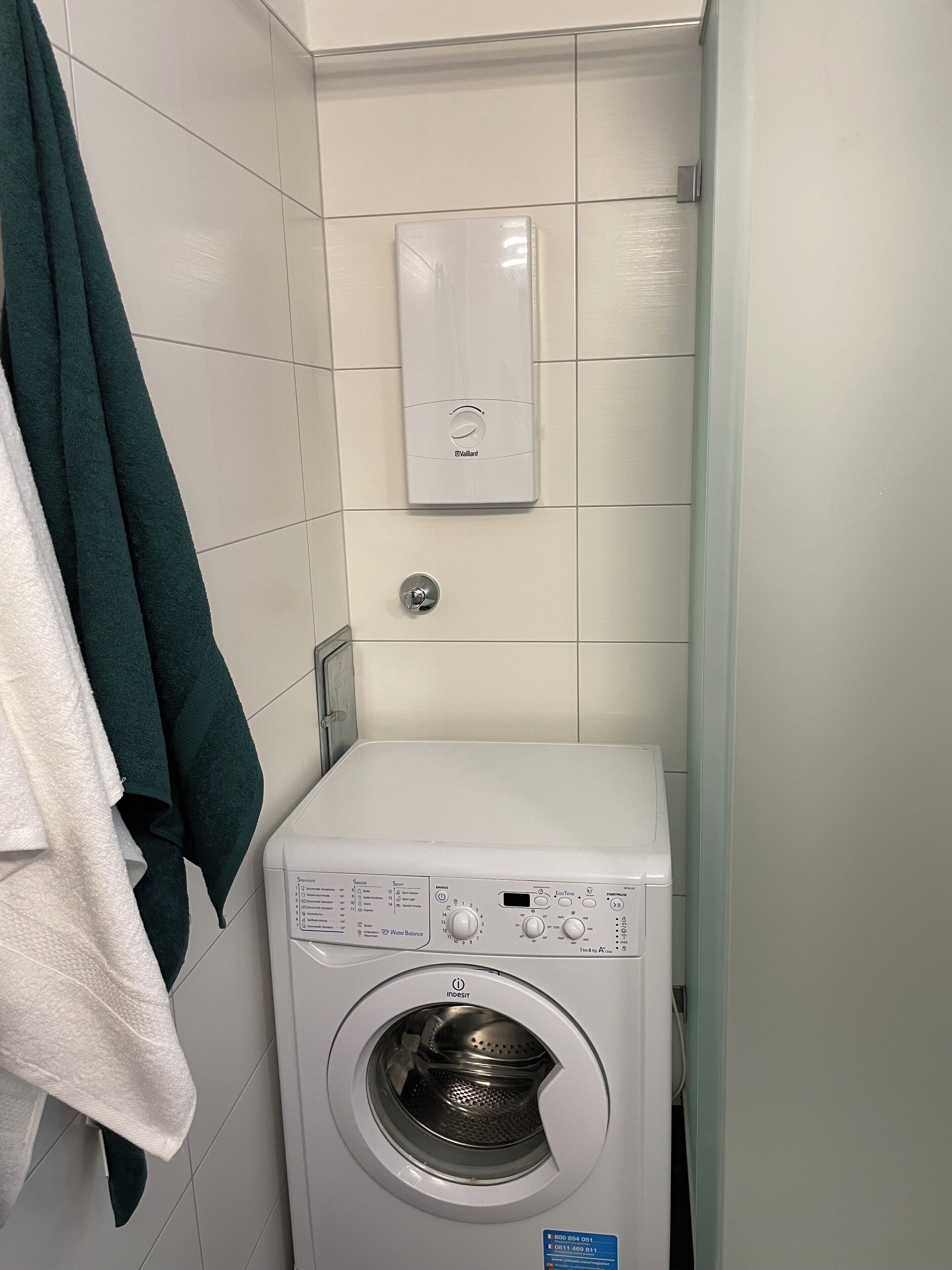 A compact, white, front-loading washing machine in a small laundry room with tiled walls and a towel rack.