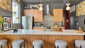 Une cuisine moderne avec un plan de travail blanc, des armoires en bois et une hotte de cuisine en acier inoxydable. On y trouve quatre tabourets de bar gris et une tête d’animal décorative accrochée au mur.