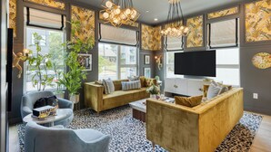 Un salon moderne avec un canapé, des fauteuils et une table basse. De grandes fenêtres sont équipées de stores, et on y trouve également un téléviseur et des œuvres d'art décoratives accrochées au mur.