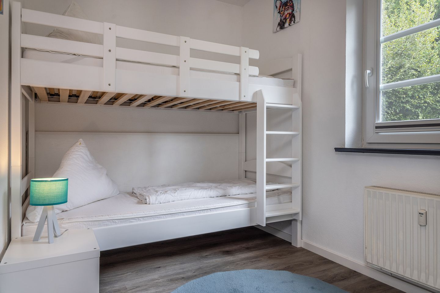 A bunk bed room with a window, a radiator, and a lamp on a bedside table.