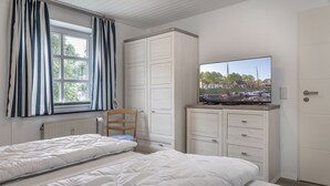 Une chambre avec deux lits, une chaise en bois, un téléviseur posé sur un meuble et une fenêtre avec des rideaux.
