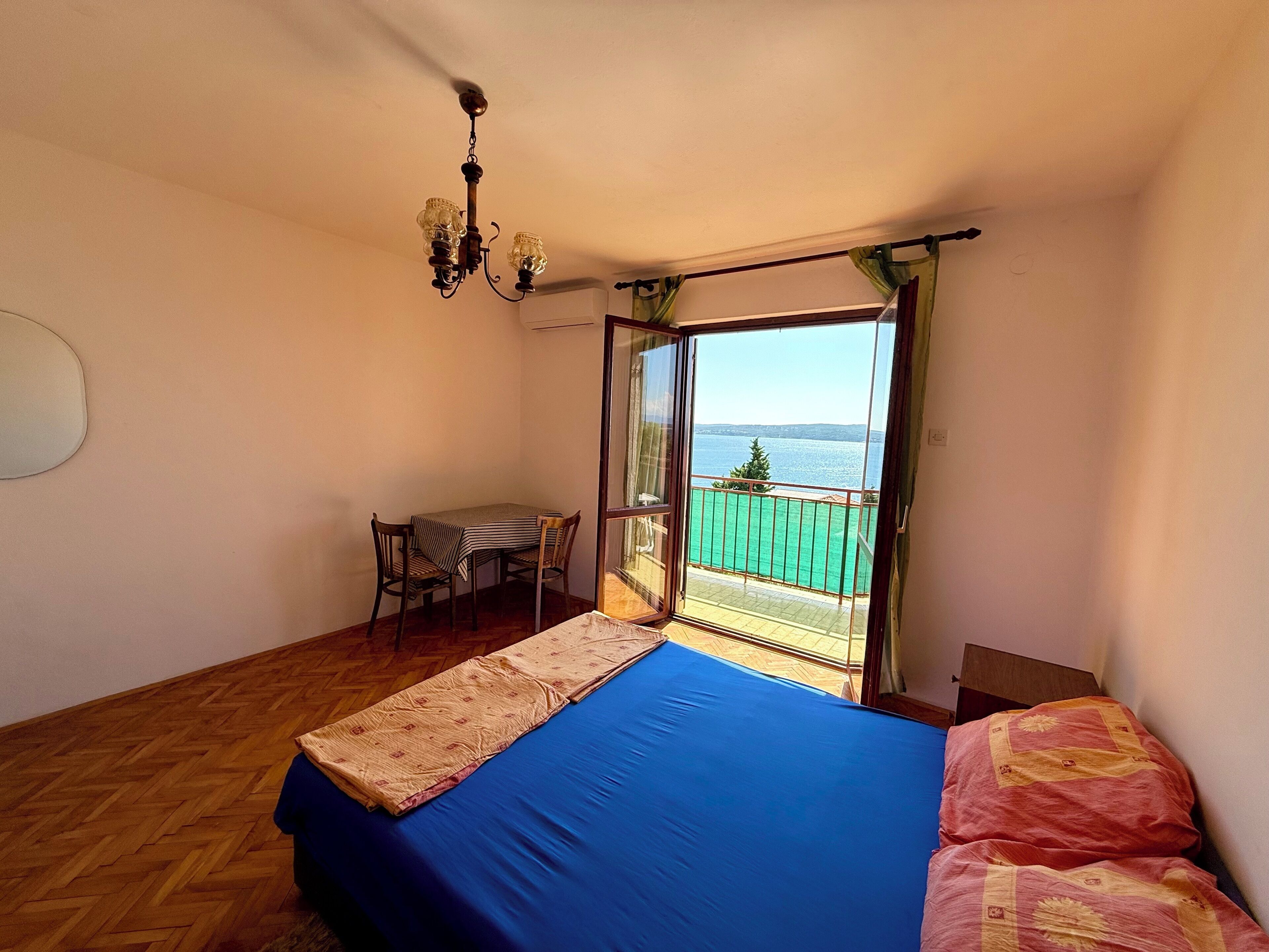 A bedroom with a bed, a small table, and a chair. There is a balcony with a view of the sea.