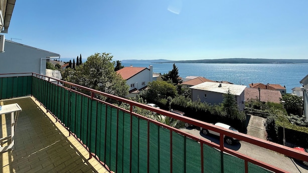 Un balcón con barandilla verde, con vistas a una zona costera con casas y un cielo azul despejado.
