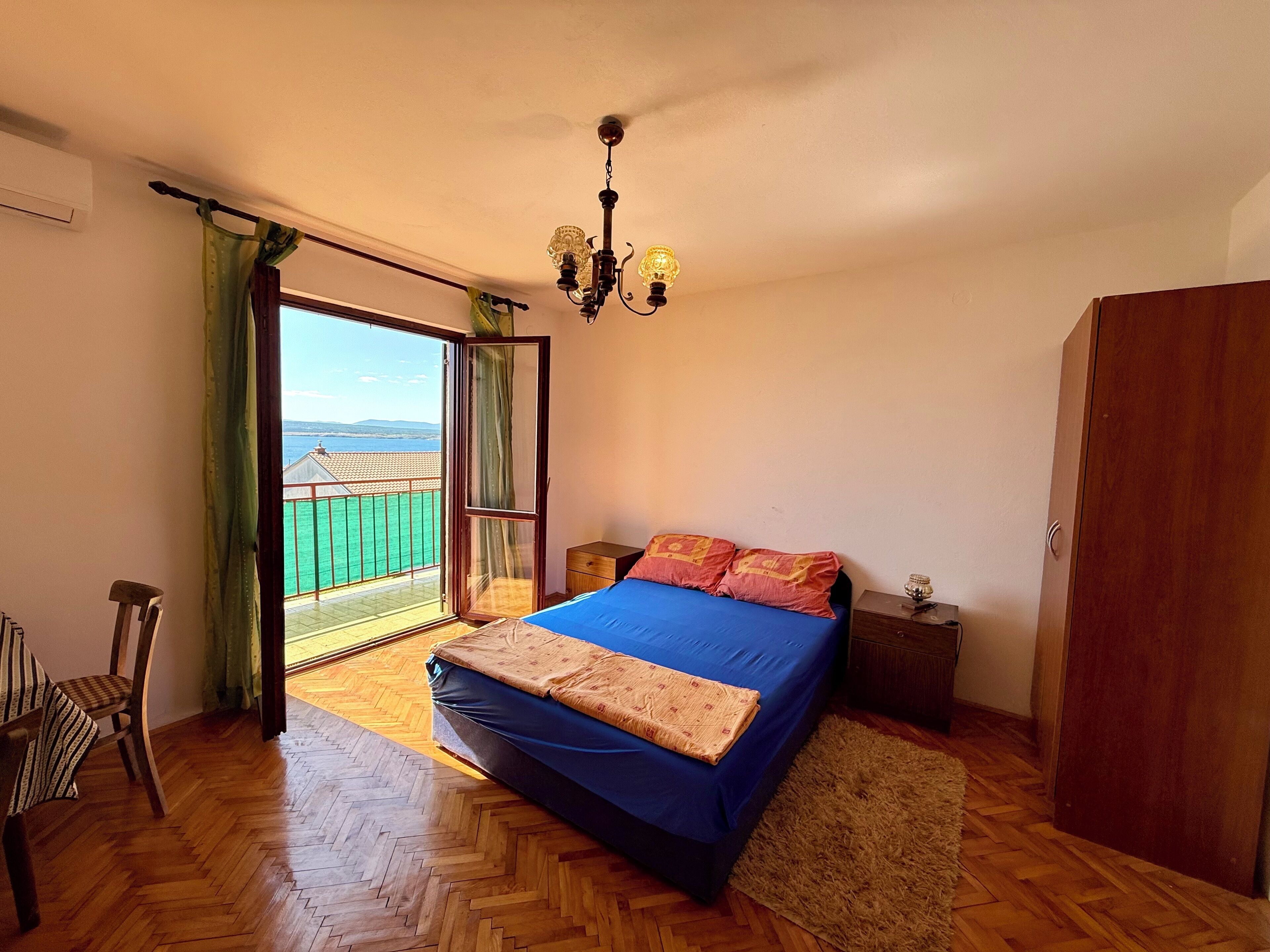Un dormitorio con cama, suelo de madera, mesita de noche, una silla, armario y balcón con vistas al mar.