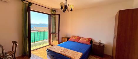 Un dormitorio con cama, suelo de madera, mesita de noche, una silla, armario y balcón con vistas al mar.