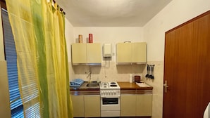 Cocina compacta con estufa a gas, lavamanos y armarios. Hay una ventana con persianas y cortina.