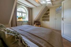 A bedroom with a large bed, a desk, a chair, a wardrobe, and a window with a view of a dog.