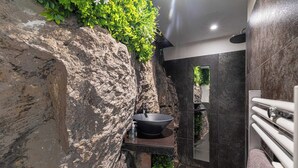 Une salle de bain avec un lavabo noir, une douche et un mur en pierre agrémenté de plantes.