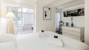 Une chambre d’hôtel comprenant un lit, une table de chevet, une lampe, une décoration murale représentant une jeune fille avec un cœur, une commode et une vue sur un balcon.