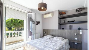 A modern bedroom with a bed, a ceiling light, a wall-mounted air conditioner, and a balcony with a view of trees.