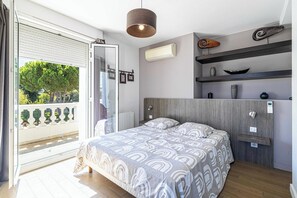 A modern bedroom with a bed, a ceiling light, a wall-mounted air conditioner, and a balcony with a view of trees.