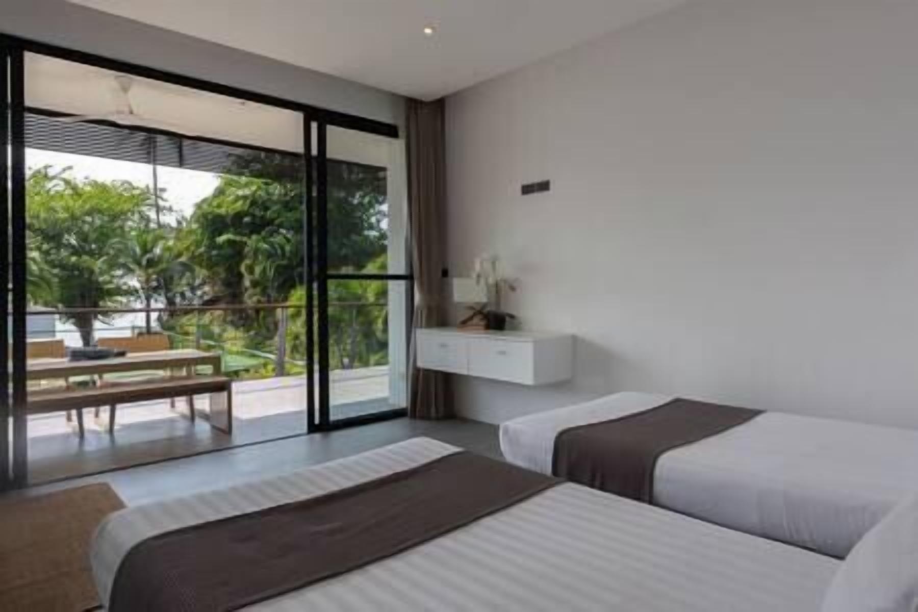 Una habitación de hotel moderna con dos camas, un amplio ventanal corredero que da a un espacio exterior y una pequeña mesita de noche blanca con una lámpara.