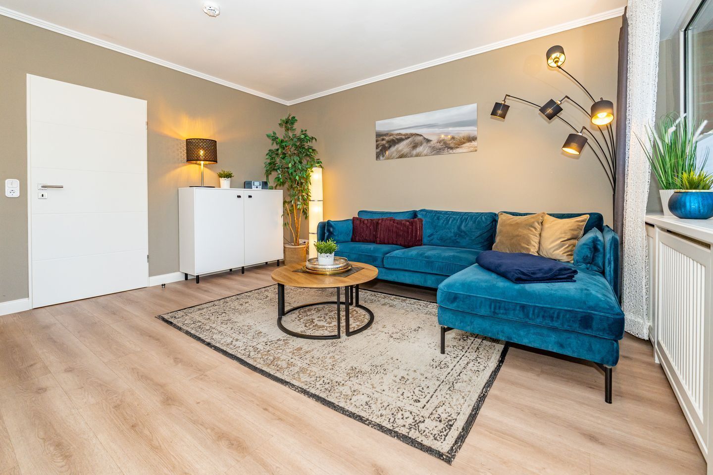 Ein modernes Wohnzimmer mit einem blauen Sofa, einem Couchtisch und einer Pflanze. Im Hintergrund sind ein weißes Schränkchen und eine Tür zu sehen.