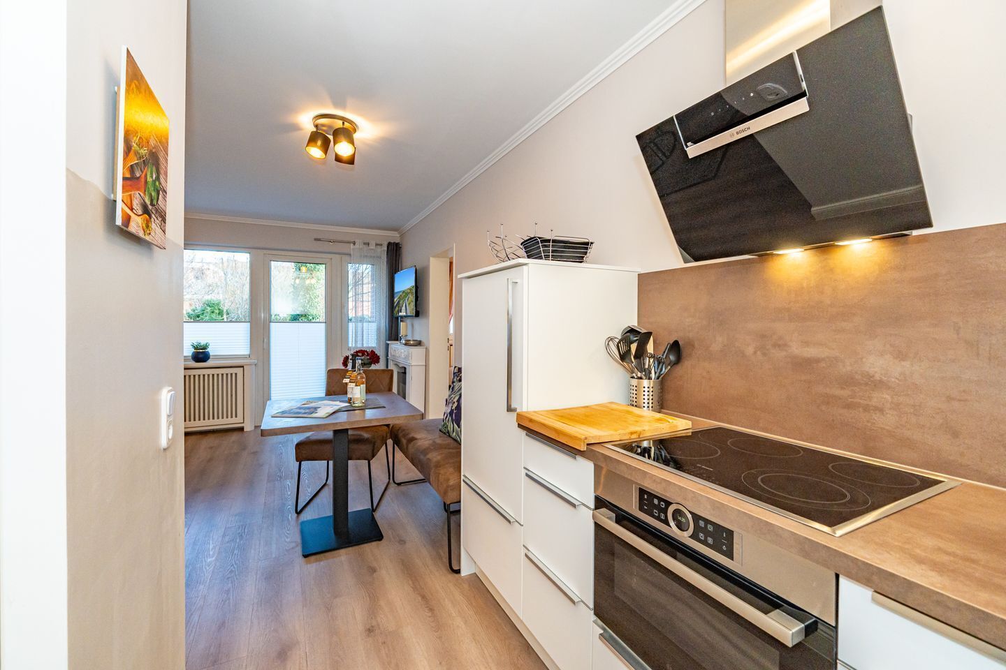 A modern kitchen with a built-in stove, oven, and white cabinetry. There is a dining area with a table and chairs, and a window providing natural light.