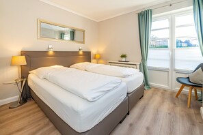 A hotel room with a double bed, bedside lamps, a small table with a plant, a chair, and a large window with curtains.