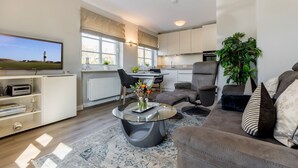 A modern living room with a flat-screen TV, a sofa, a coffee table, and a dining area with a kitchen in the background.
