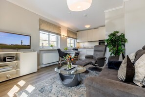 A modern living room with a flat-screen TV, a sofa, a coffee table, and a dining area with a kitchen in the background. - Syltliebe - Holiday apartment with terrace and garden close to the beach on Sylt (Sylt)