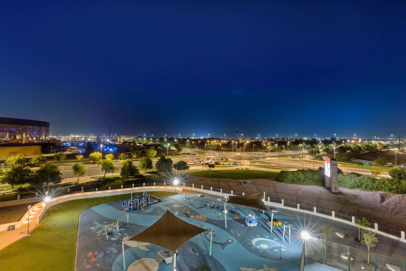 A nighttime view of a well-lit park with a playground, a large building with lights in the background, and a clear sky.