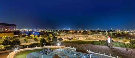 A nighttime view of a well-lit park with a playground, a large building with lights in the background, and a clear sky.