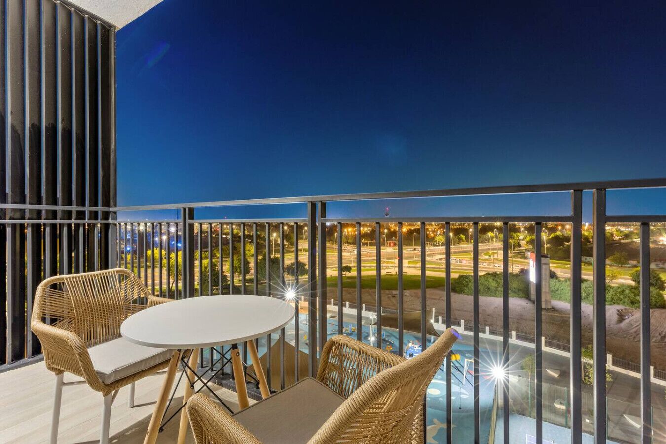 A balcony with wicker furniture, a glass table, and a view of a cityscape at night.