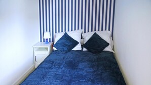 Un lit recouvert d’une couverture bleu foncé à texture marquée, deux oreillers, une table de chevet blanche avec une lampe, et un mur à rayures.